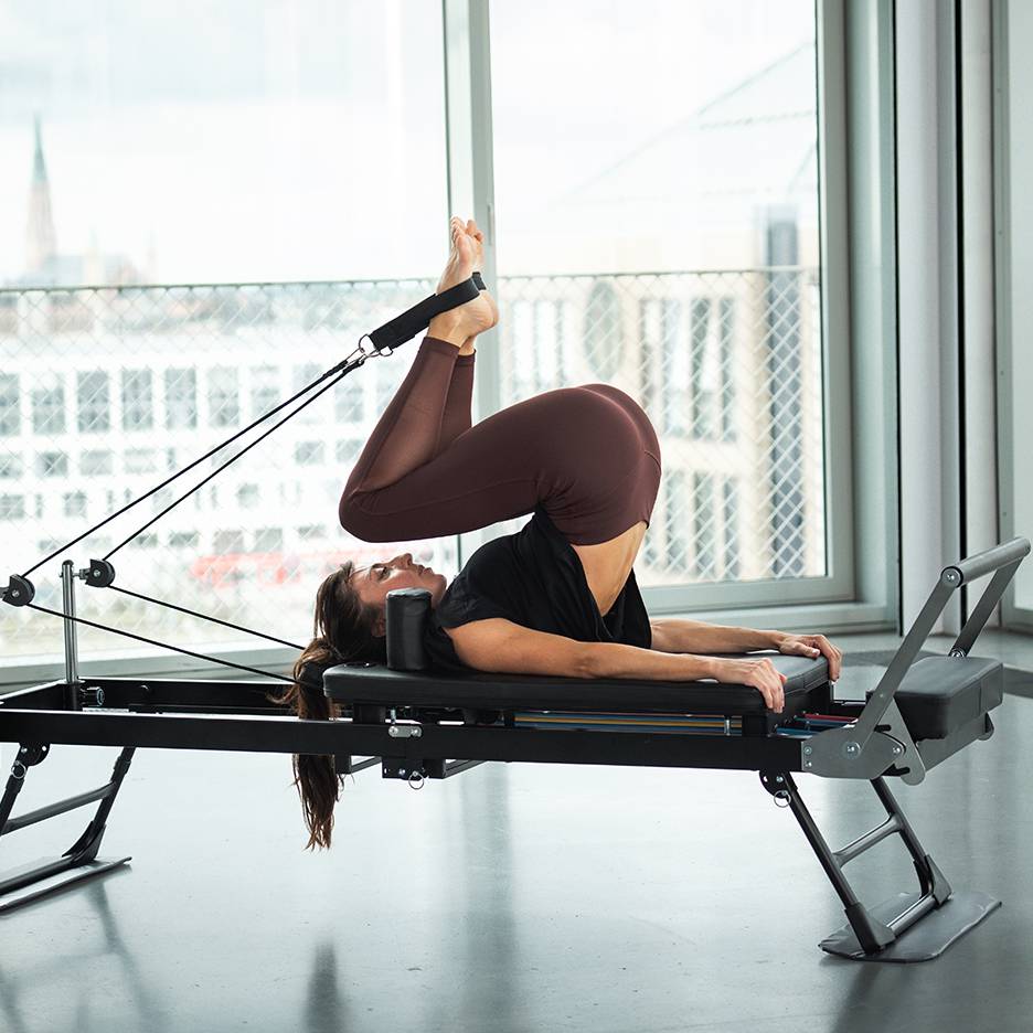 Žena cvičí Pilates na reformer stroji s nohami hore a chodidlami v popruhoch.