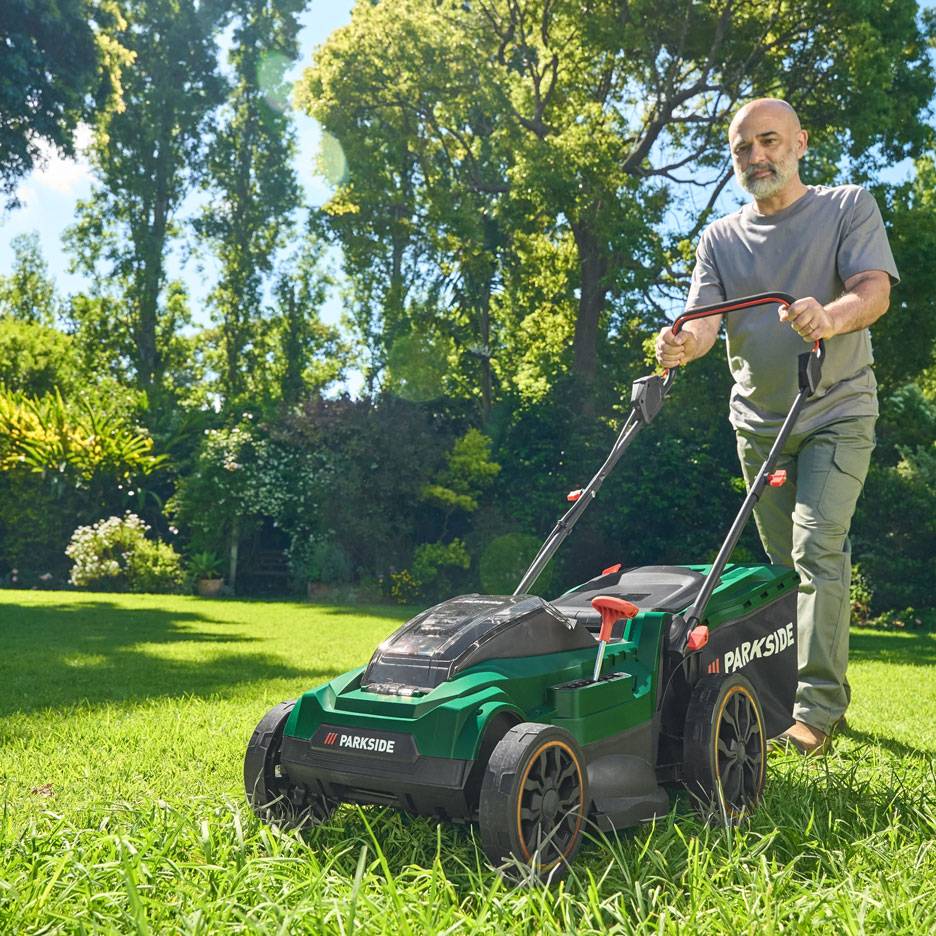 Muž kosí trávu zelenou kosačkou v slnečnej záhrade.