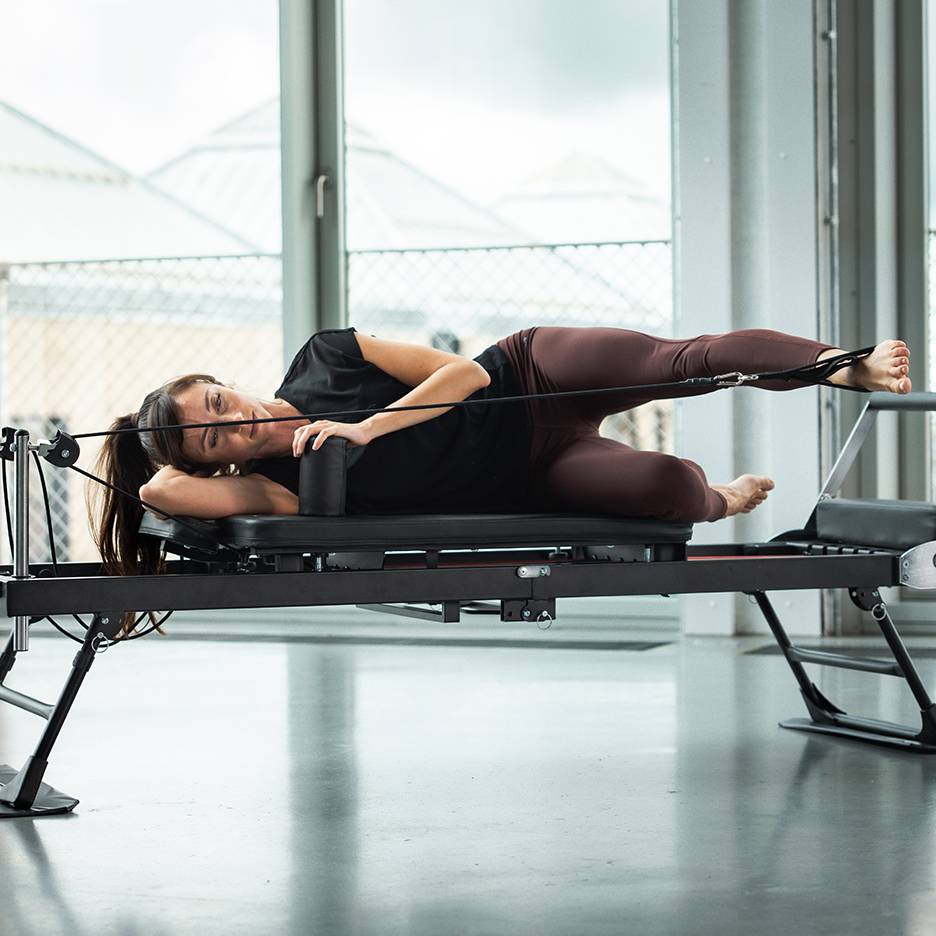 Žena cvičí Pilates na reformer stroji, ležiaca na boku s popruhmi na nohy.