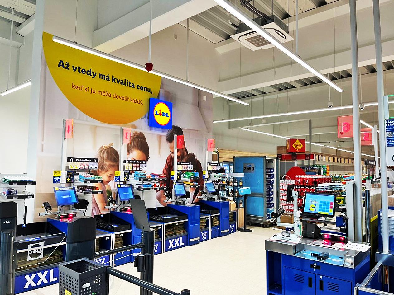 Samoobslužné pokladne v supermarkete s nápisom „Až vtedy má kvalita cenu, keď si ju môže dovoliť každý.“