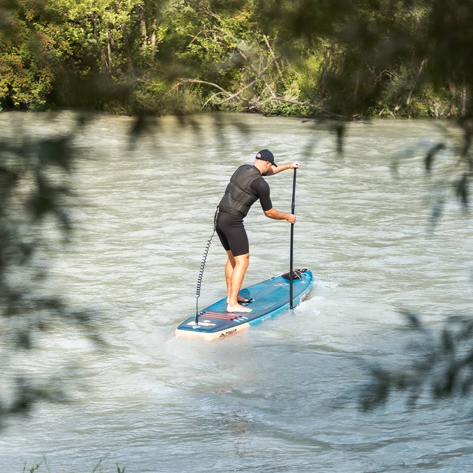 Muž v neoprénovom obleku a záchrannej veste pádluje na modrom paddleboarde na rieke.