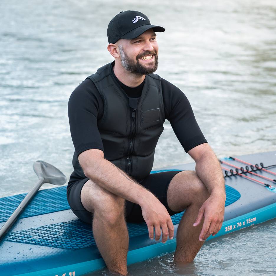 Usmievajúci sa muž v záchrannej veste a čiapke, sediaci na paddleboarde vo vode.