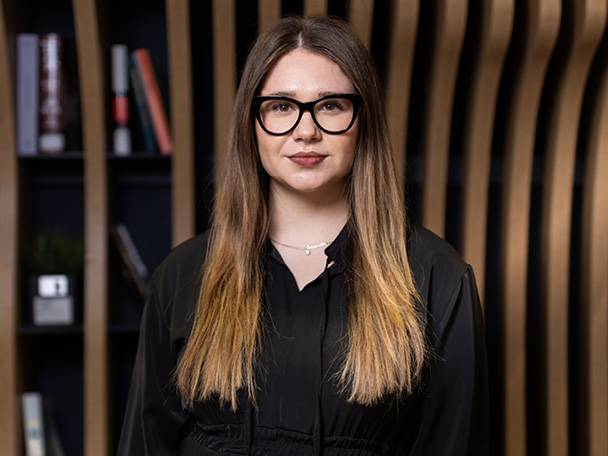 Žena s okuliarmi a dlhými vlasmi v čiernej blúzke, s knižnicou v pozadí.