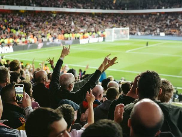 Fanúšikovia na futbalovom štadióne, niektorí s rukami vo vzduchu, jeden fotí telefónom.