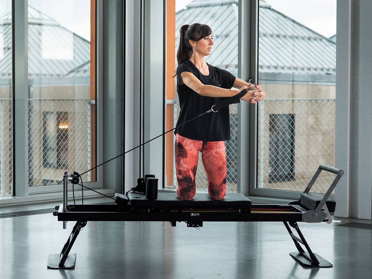 Žena cvičí Pilates na čiernom reformer stroji vo svetlej miestnosti.
