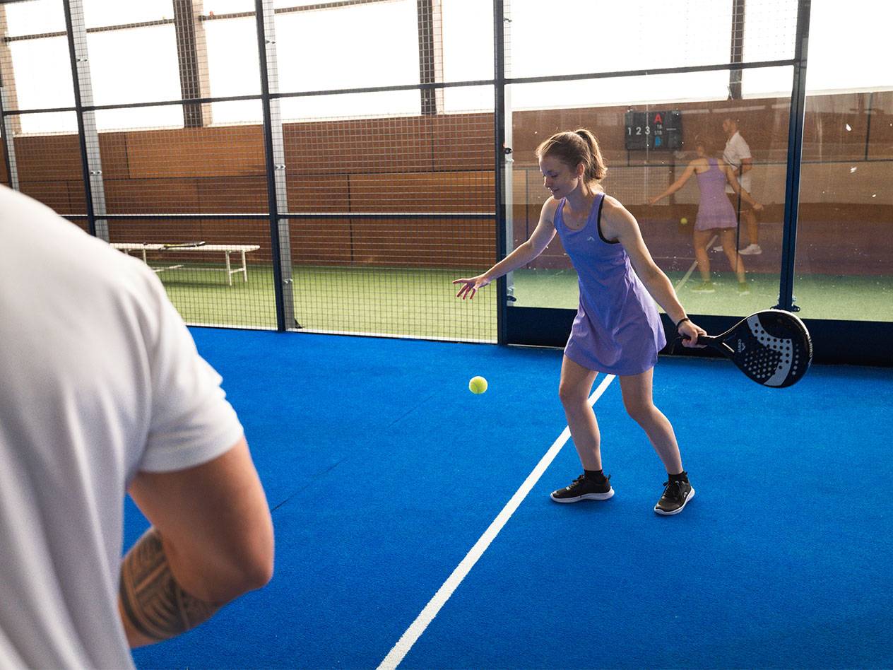 Žena hrá padel, drží raketu a pozerá na loptičku na modrom kurte.