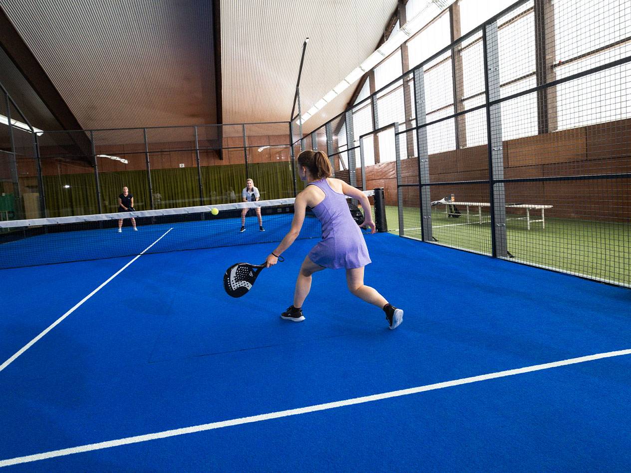 Žena hrá padel na modrom kurte, s ďalšími hráčmi v pozadí.