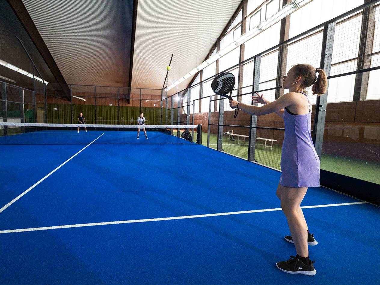 Žena vo fialových šatách hrá padel na modrom kurte, ostatní hráči v pozadí.