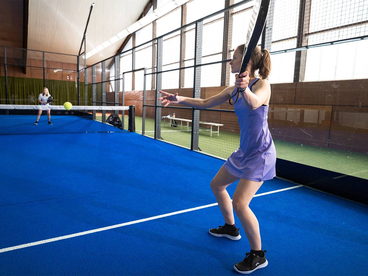 Žena vo fialových športových šatách hrá padel na modrom kurte.