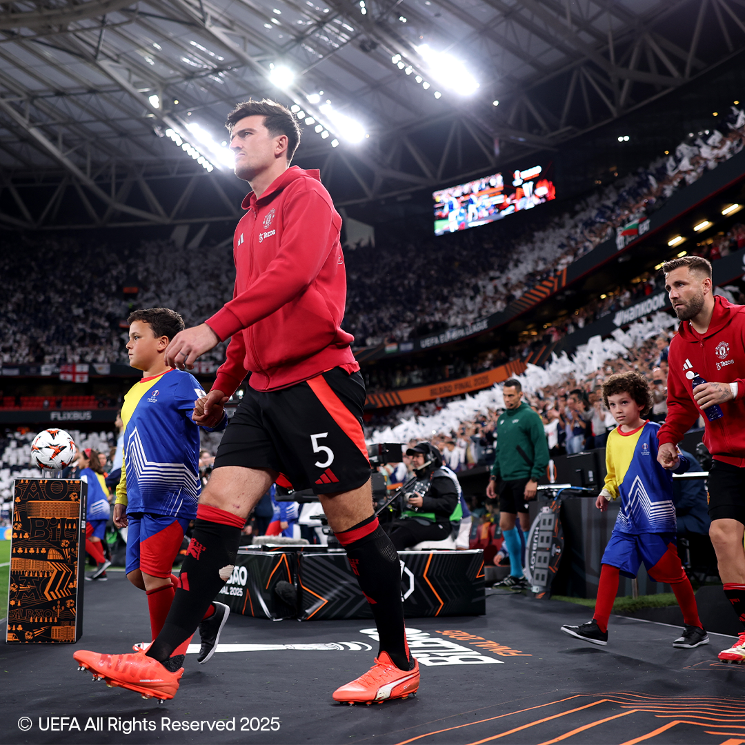 Futbalisti v červených dresoch a čiernych šortkách kráčajú po ihrisku s deťmi v športovom oblečení.