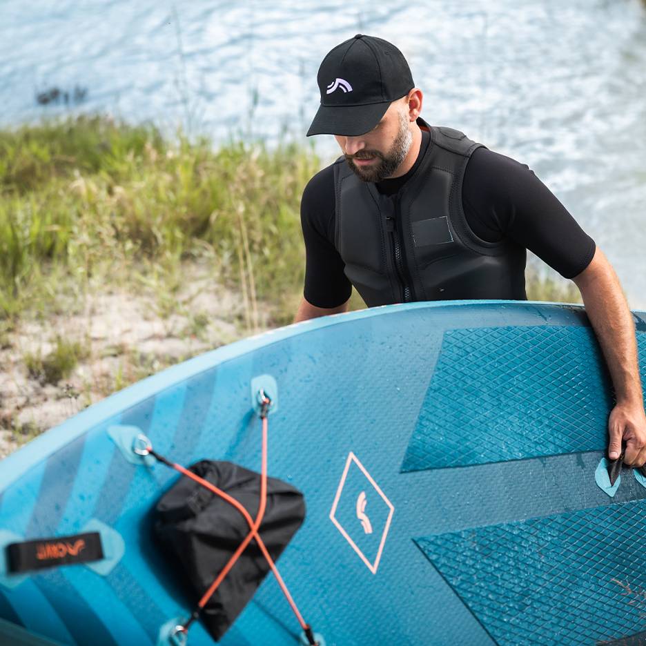 Muž v záchrannej veste a čiapke drží modrý paddleboard pri vode.