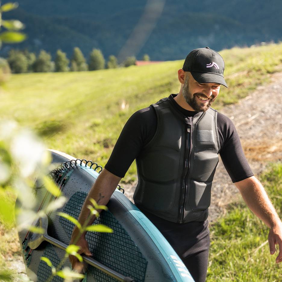 Muž v čiernom neopréne a záchrannej veste nesie paddleboard s pádlom.