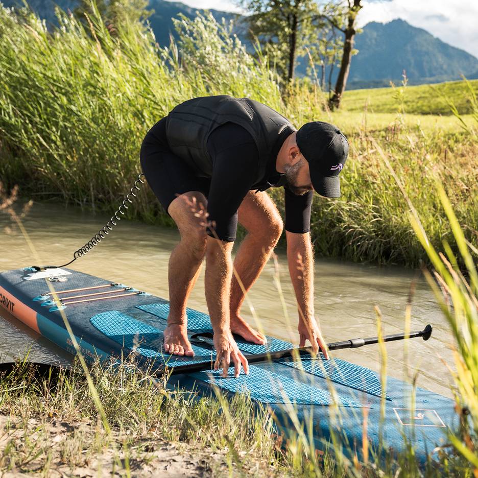 Muž v neoprénovom obleku a záchrannej veste sa pripravuje na paddleboarding na rieke.