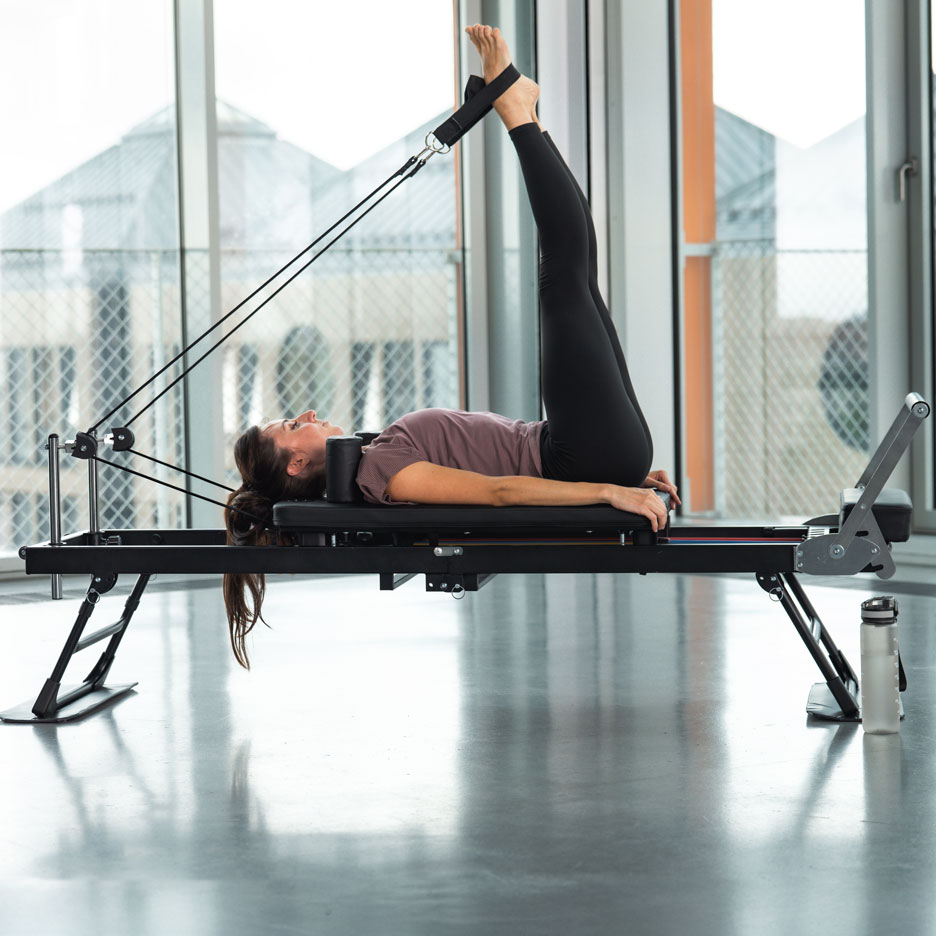 Žena cvičí Pilates na reformer stroji s nohami hore, vo svetlej miestnosti.
