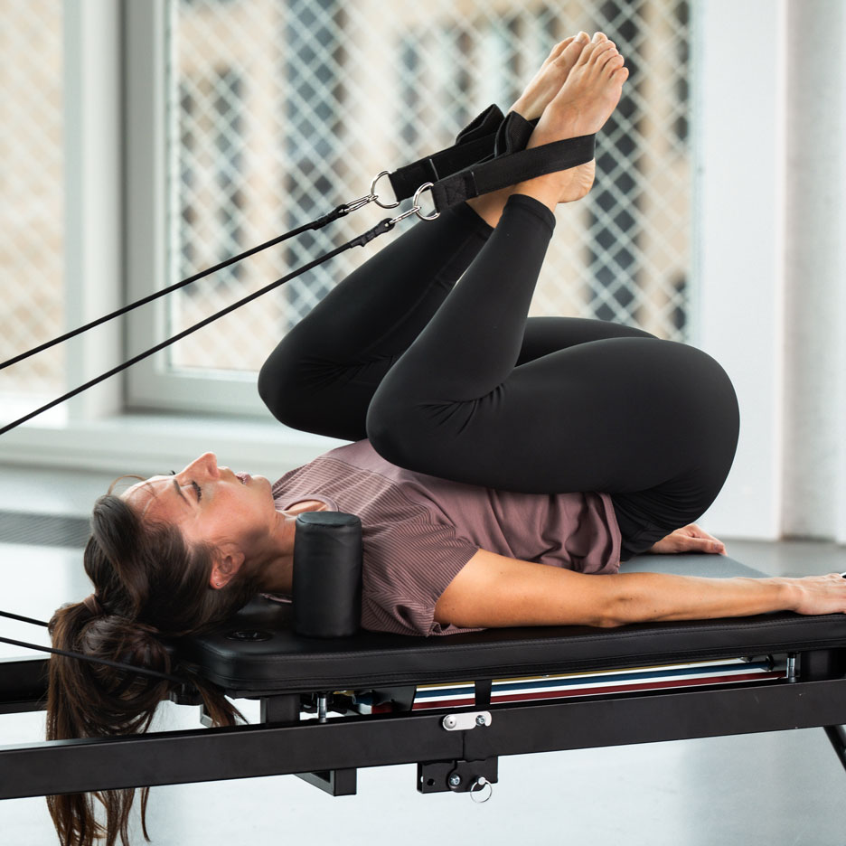 Žena cvičiaca Pilates na reformer stroji s nohami v popruhoch.