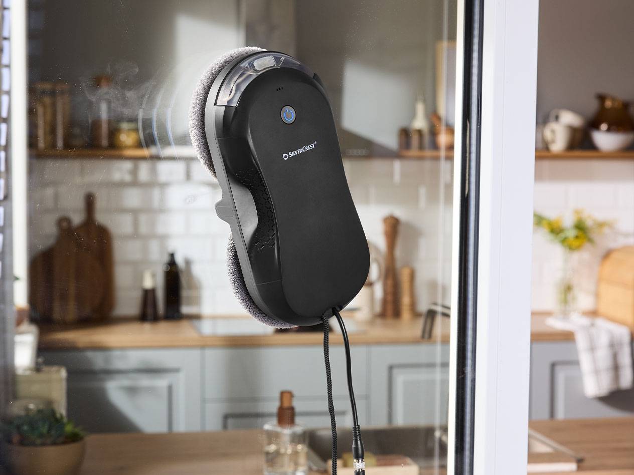 Robot na čistenie okien Silvercrest na okennom skle s kuchyňou v pozadí.