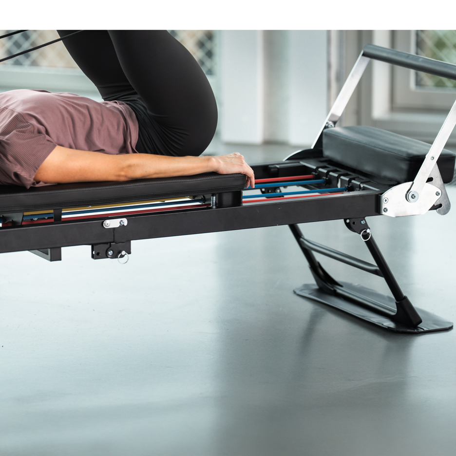 Žena cvičiaca na čiernom Pilates stroji s farebnými odporovými pásmi.