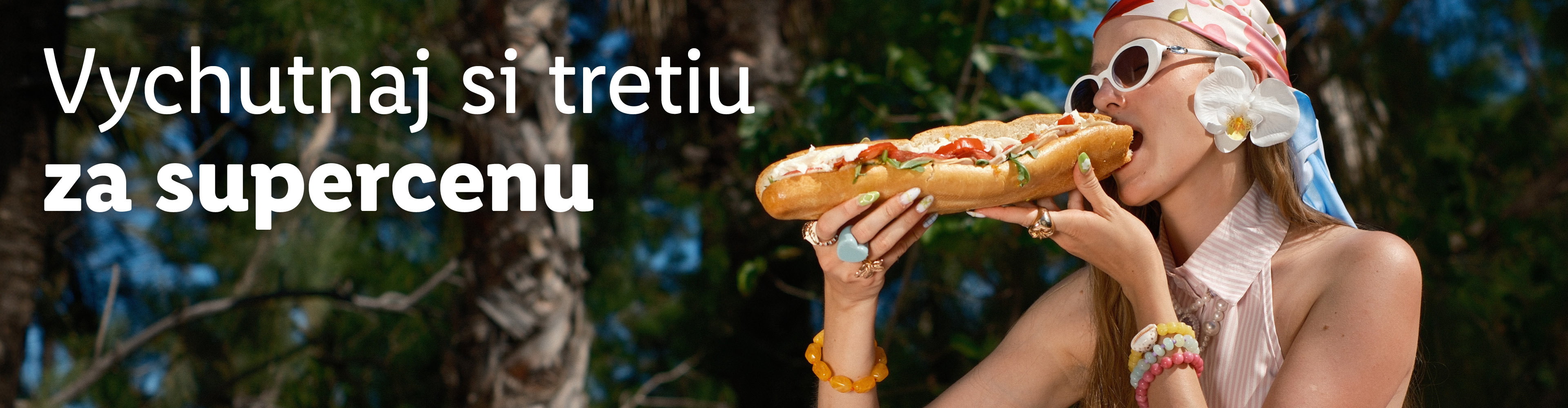 Žena s okuliarmi a šatkou na hlave jedáva dlhý sendvič. Text: Vychutnaj si tretiu za supercenu.
