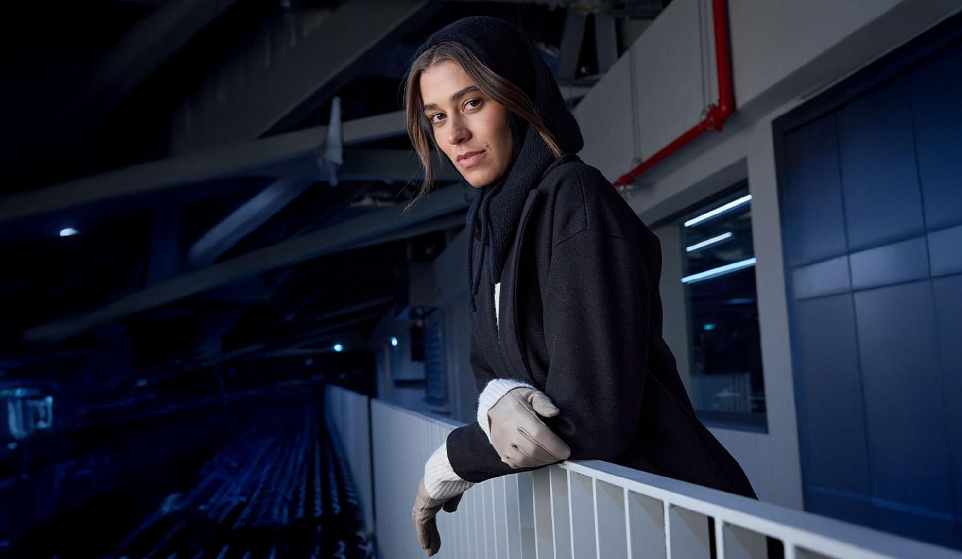 Žena v čiernom kabáte a šatke s kapucňou, opierajúca sa o zábradlie v modernom prostredí.