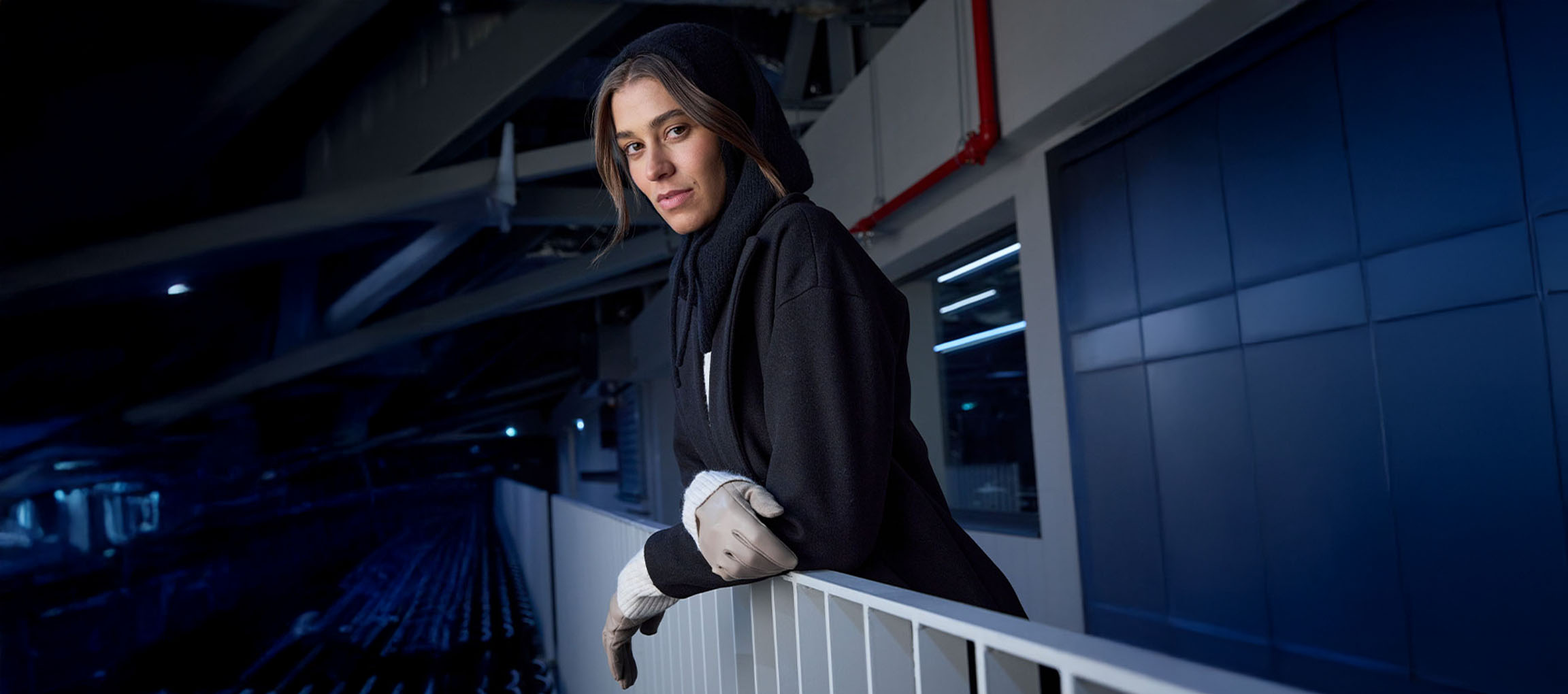 Žena v čiernom kabáte a rukaviciach opretá o zábradlie v mestskom prostredí.