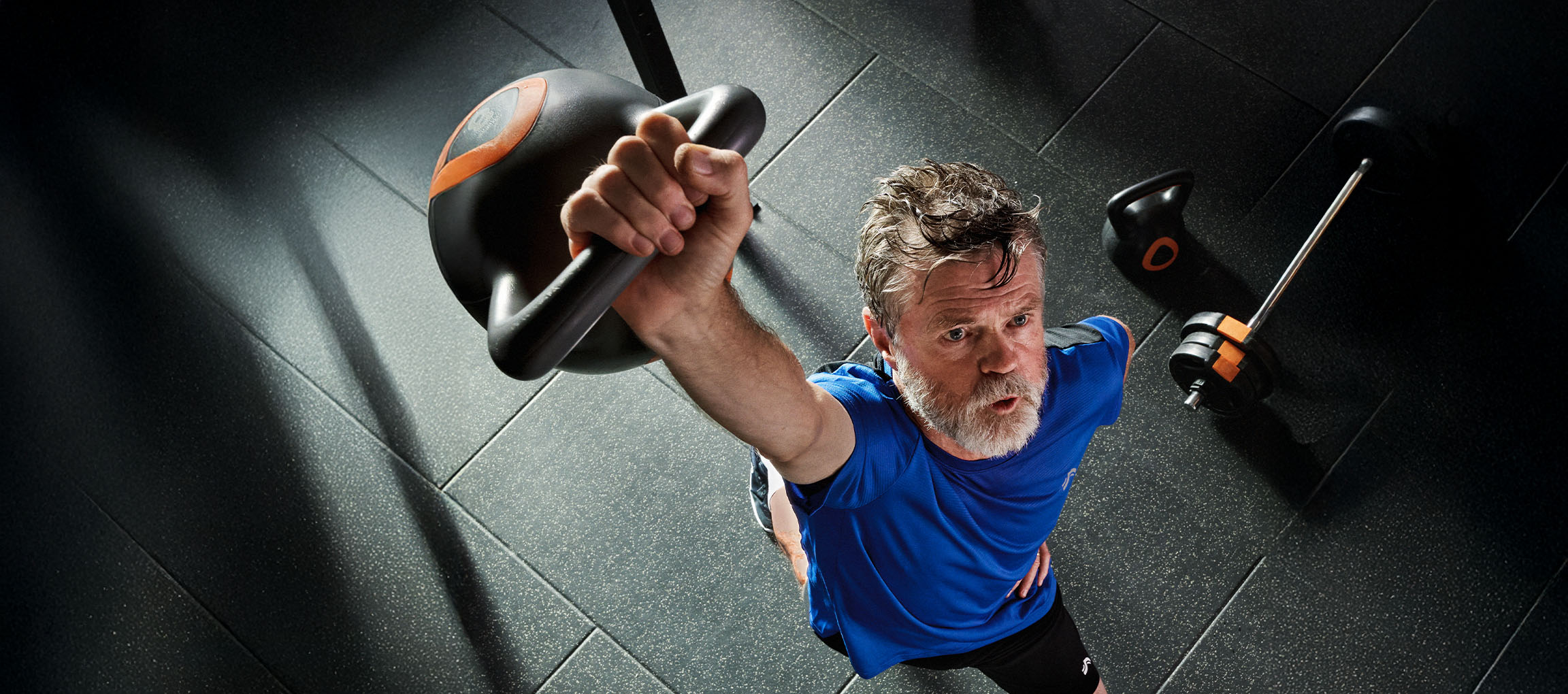 Muž dvíha kettlebell v posilňovni, s činkou a kettlebellom na podlahe.