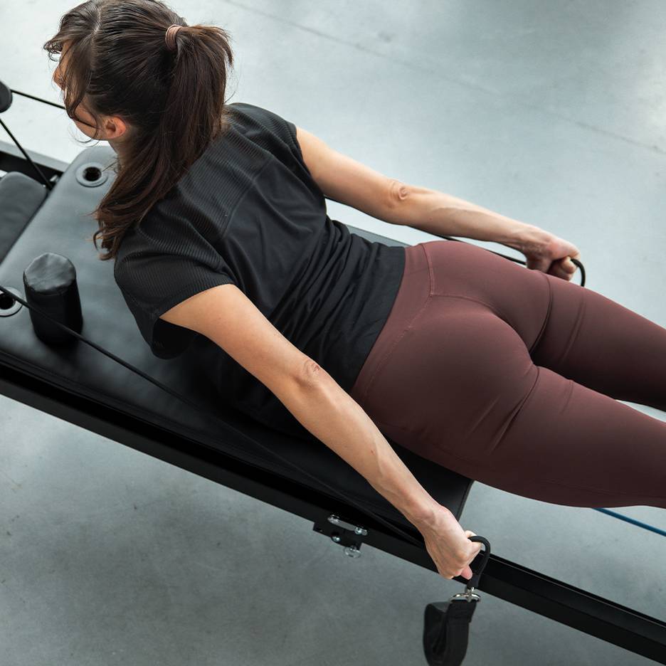Žena cvičiaca na Pilates stroji s odporovými pásmi, v športovom oblečení.