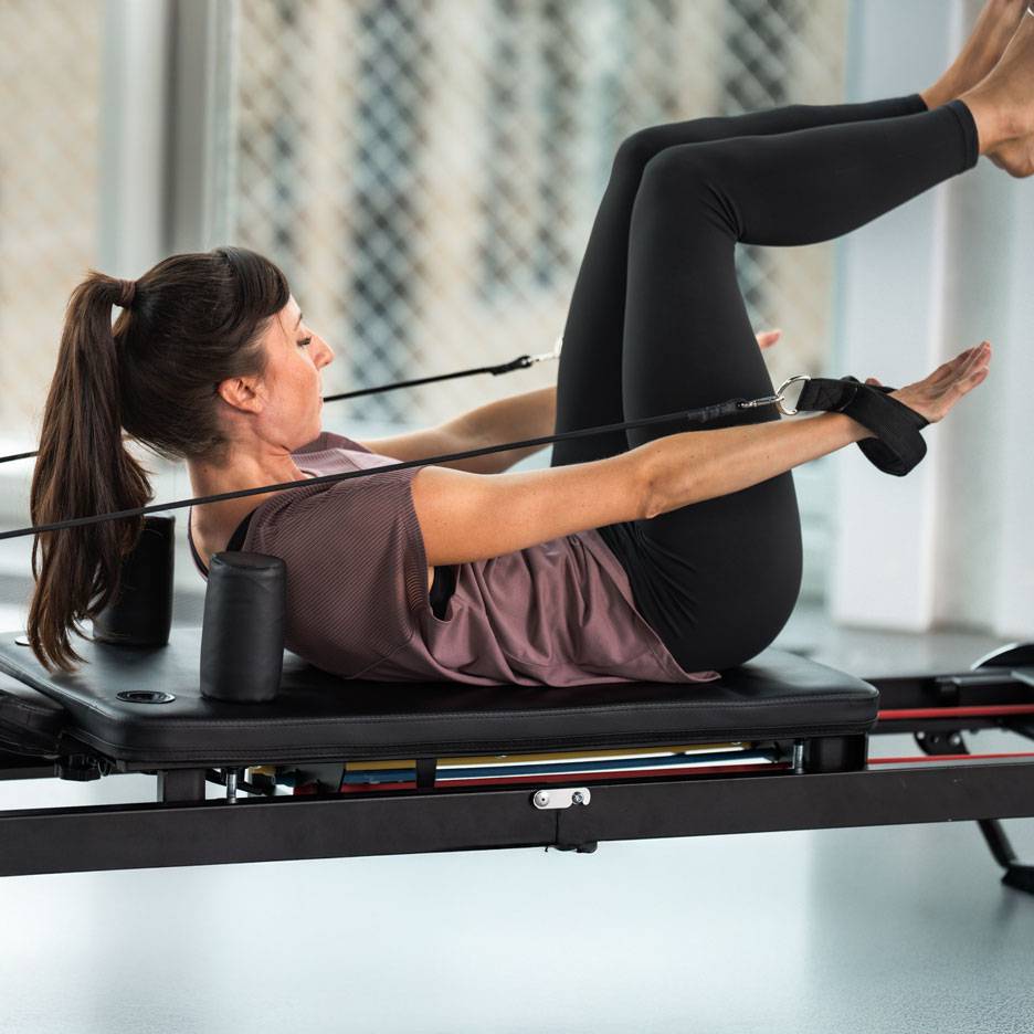 Žena cvičiaca Pilates na reformer stroji, v čiernom športovom oblečení.