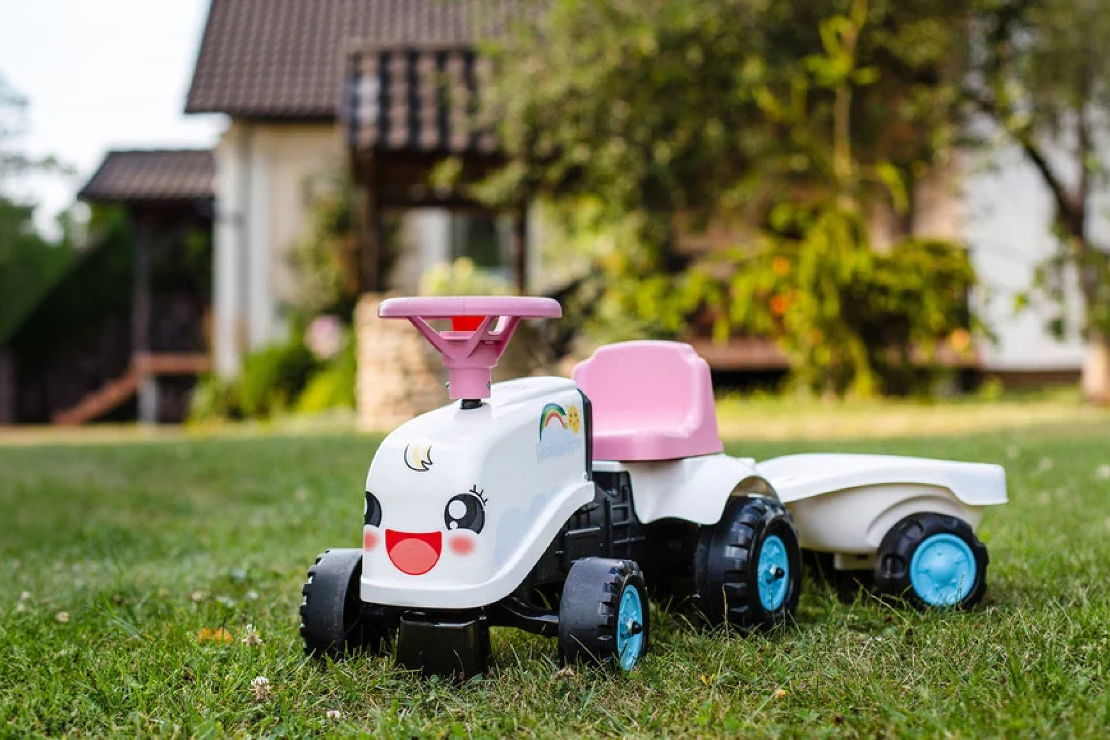 Biely detský traktor s ružovým sedadlom a prívesom na zelenej tráve pred domom.