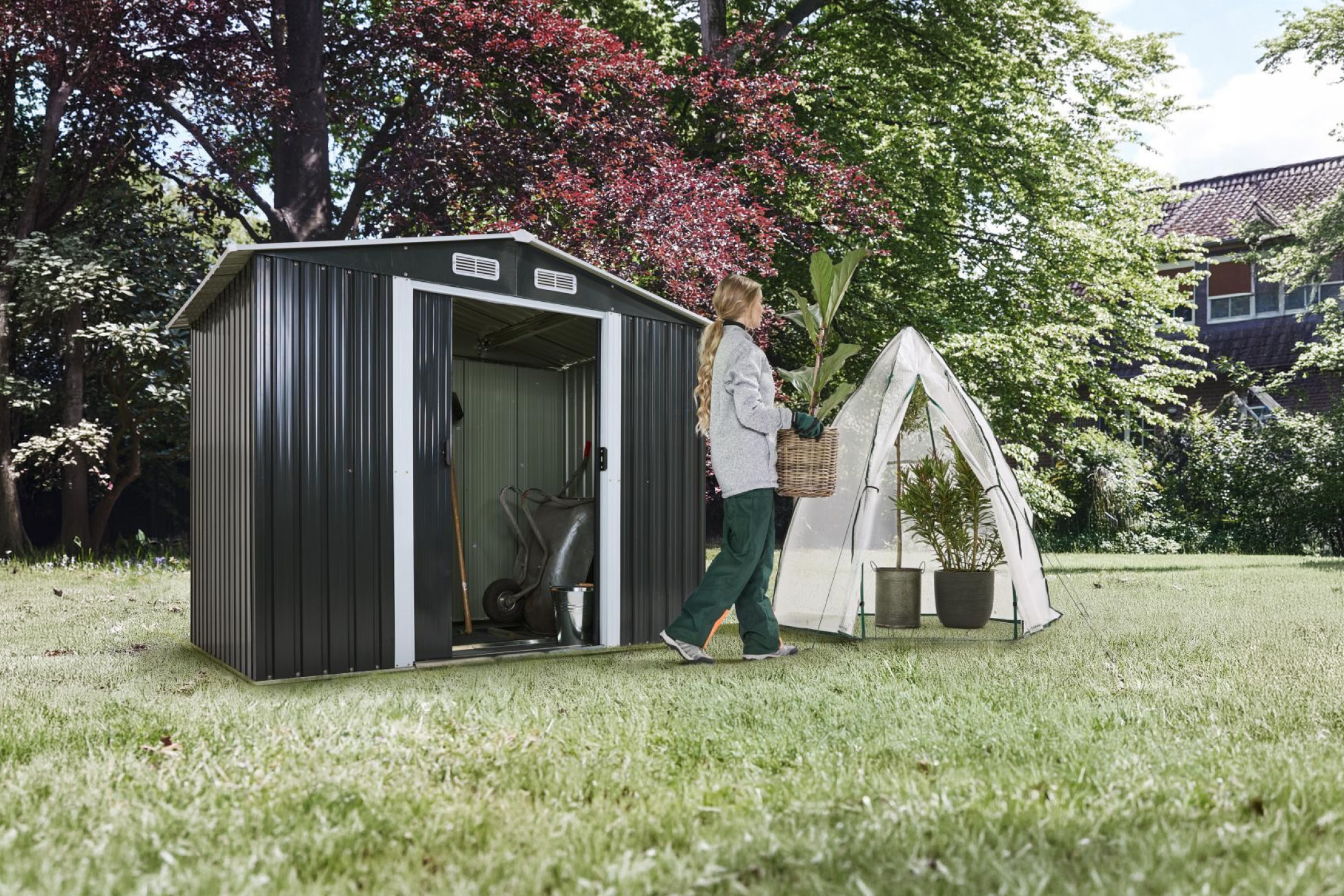 Záhradný domček a fóliovník v záhrade s osobou nesúcou rastlinu.