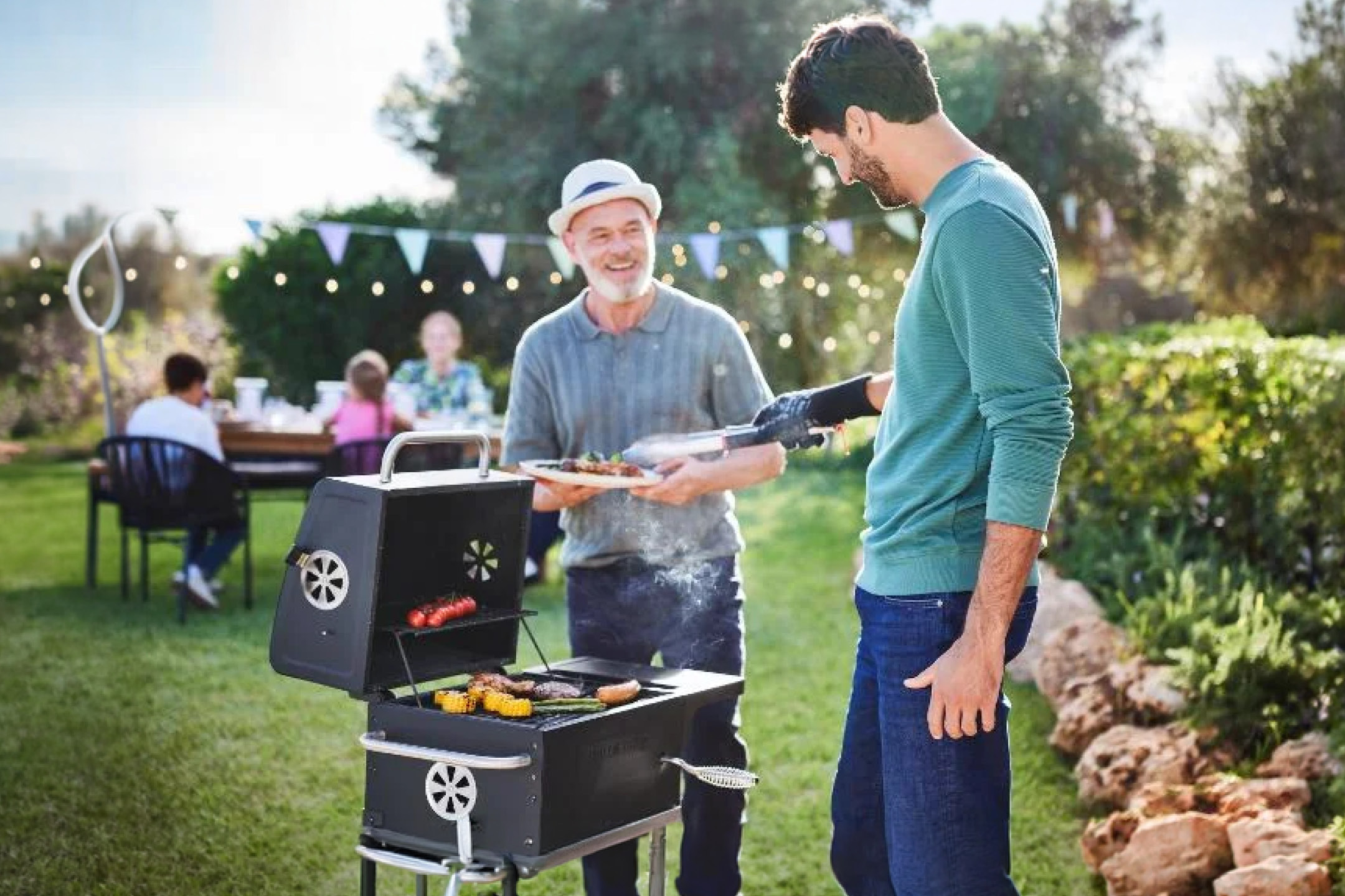 Muži grilujú mäso a zeleninu na záhradnej párty s rodinou v pozadí.