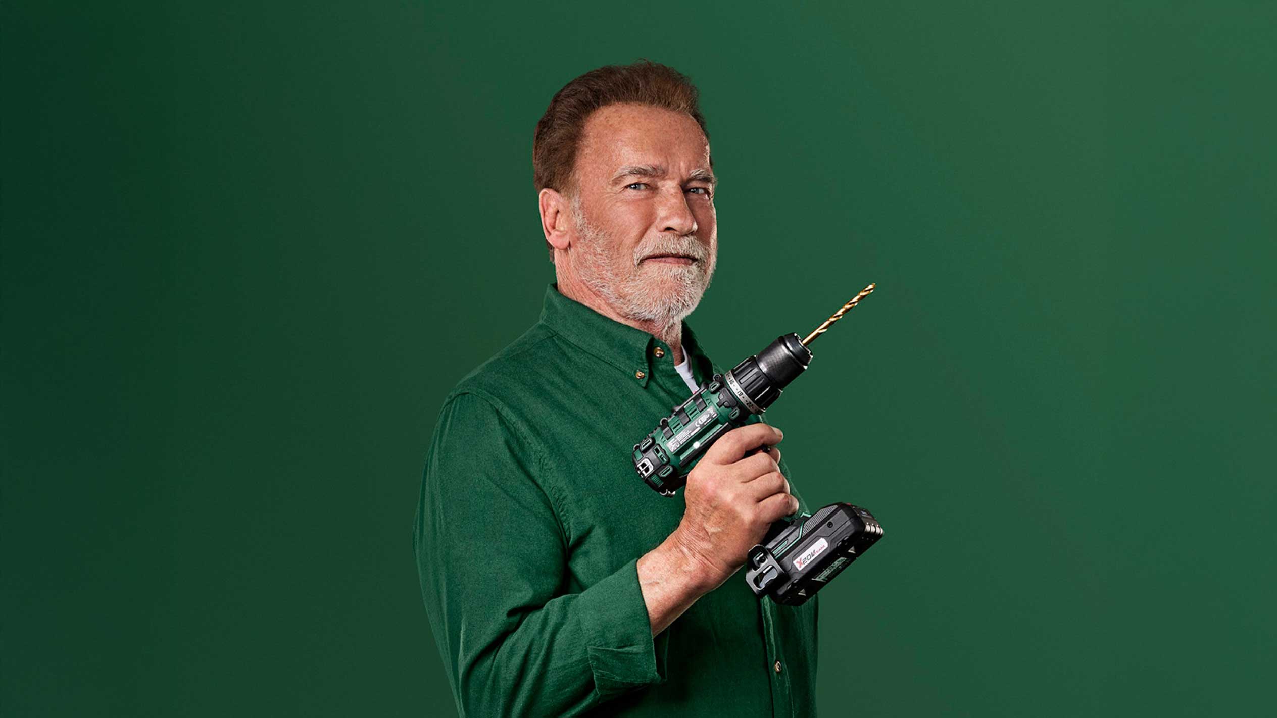 Arnold Schwarzenegger drží zelenú akumulátorovú vŕtačku so zlatým vrtákom.