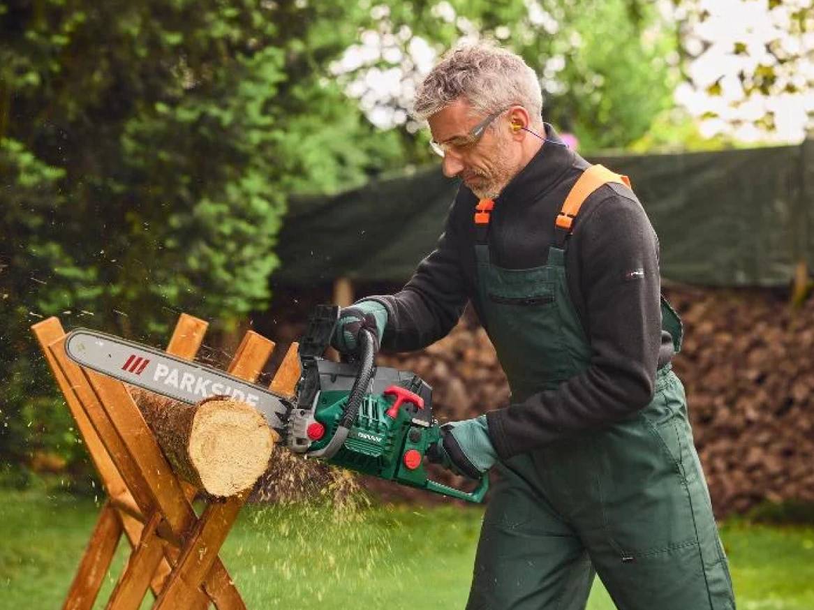Muž v montérkach a ochranných okuliaroch reže drevo reťazovou pílou na koze.