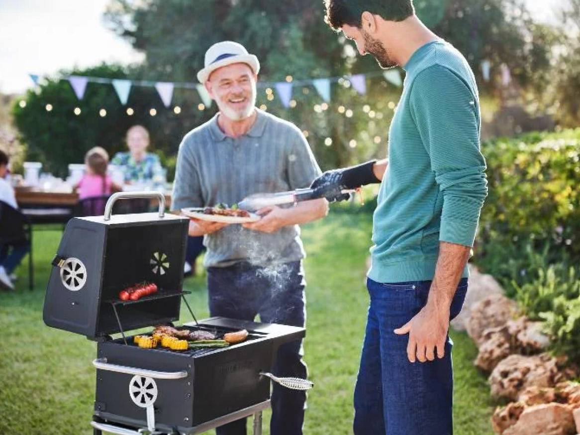 Muži grilujú v záhrade, v pozadí rodinná oslava.