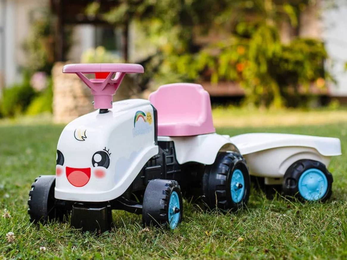 Biely detský traktor s ružovým sedadlom a volantom, s prívesom, na tráve.