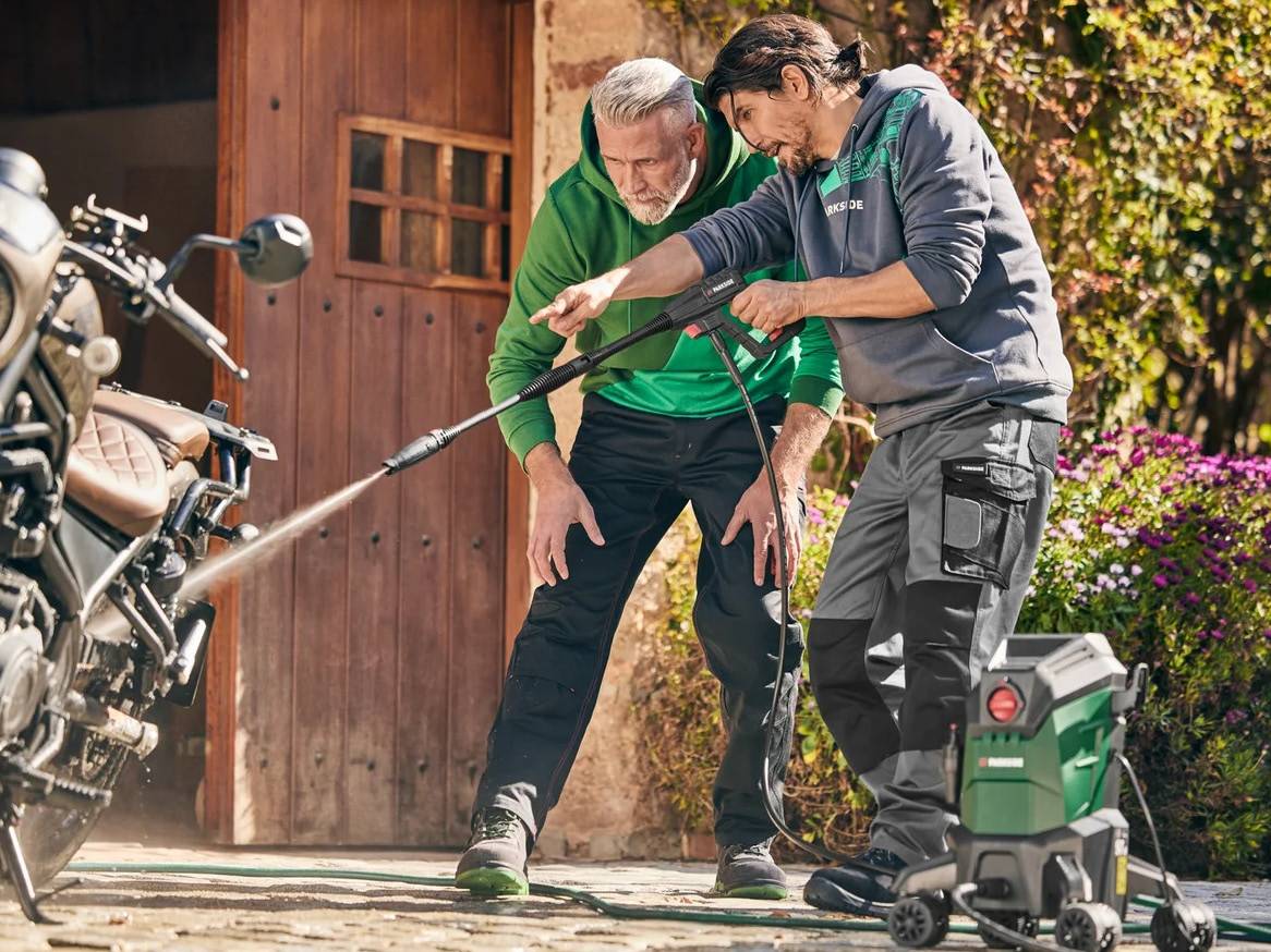 Dvaja muži umývajú motorku vysokotlakovým čističom pred drevenými dverami.