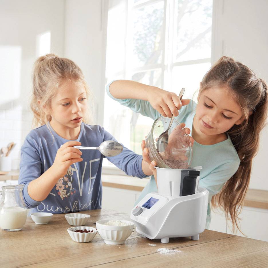Dve dievčatá pripravujú cesto v kuchynskom robote, s miskami ingrediencií na stole.