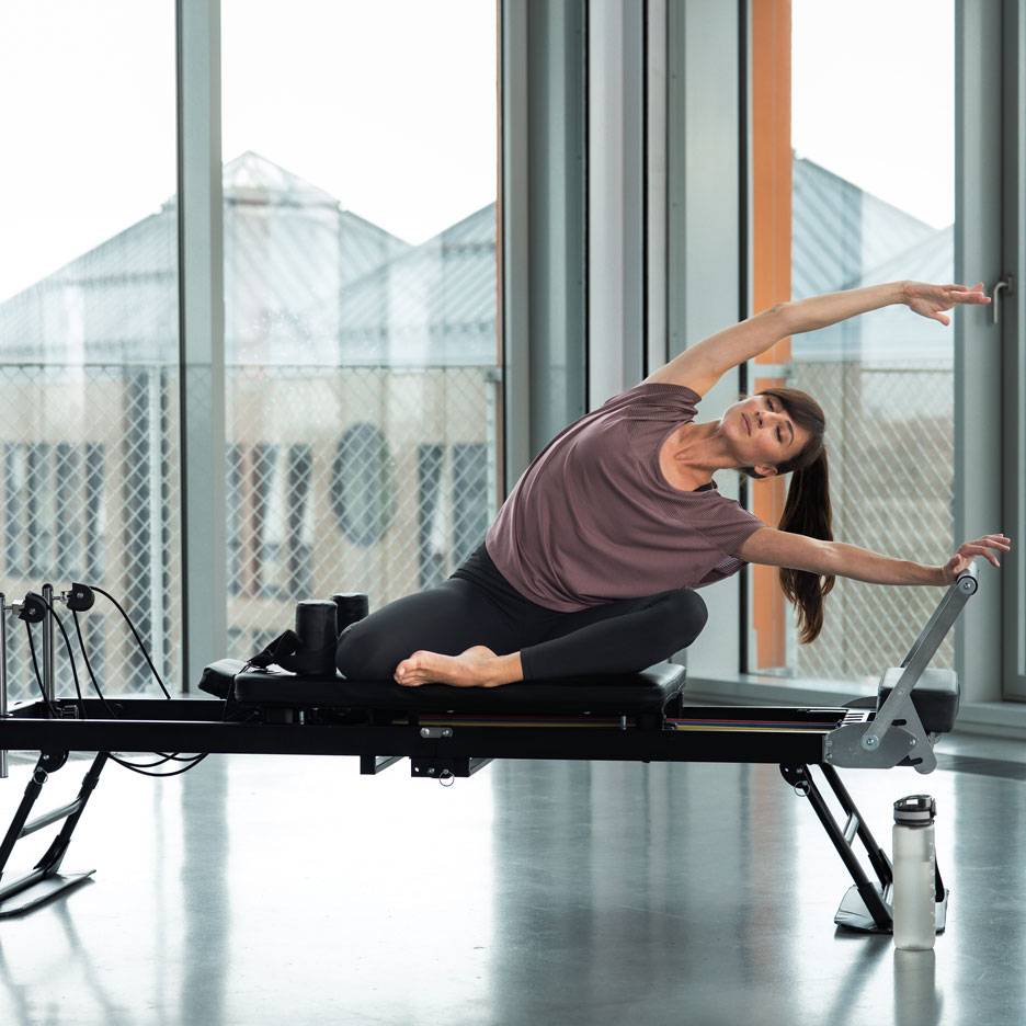 Žena cvičí pilates na reformer stroji, naťahuje sa do strany.