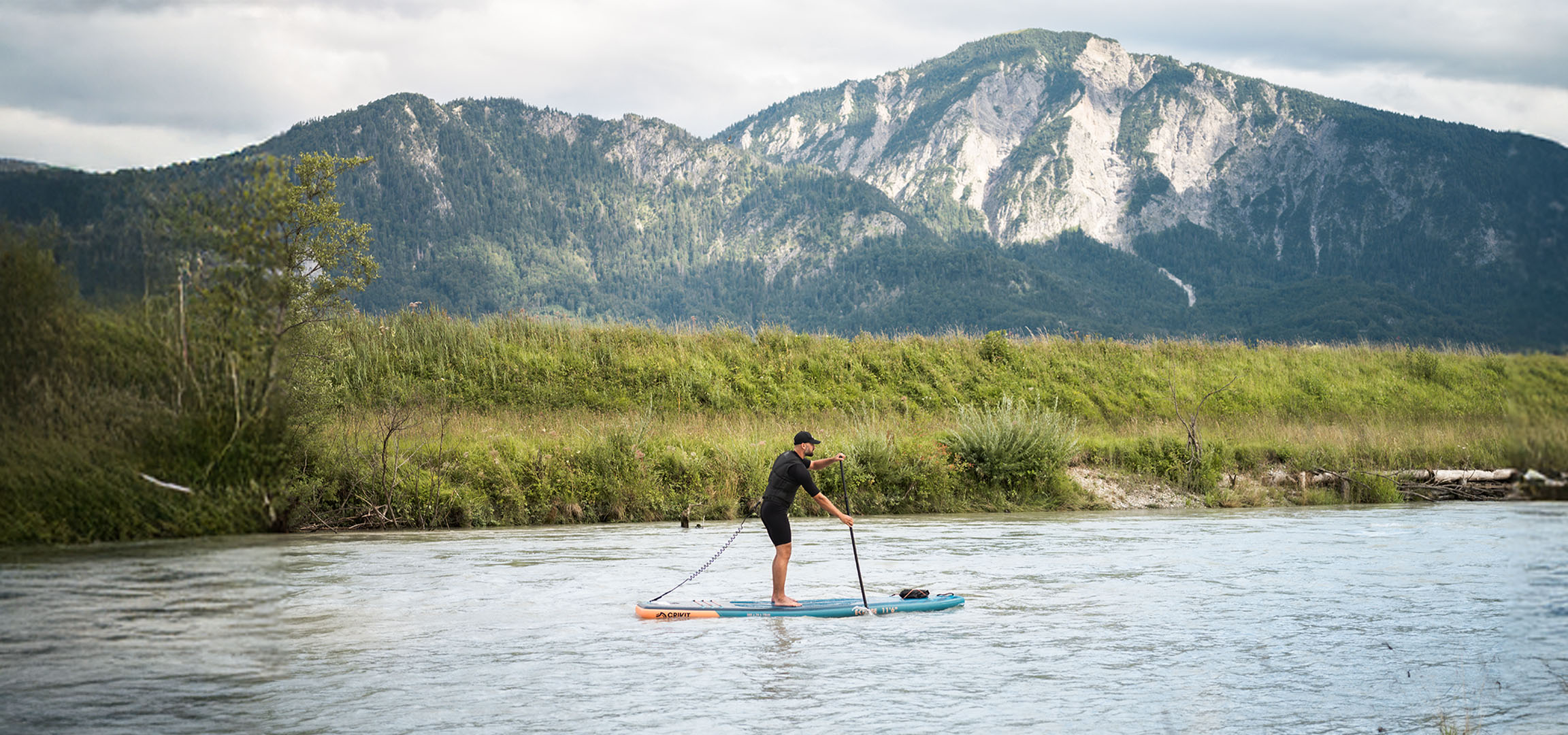 Muž na paddleboarde na rieke s horami a bujnou vegetáciou v pozadí.