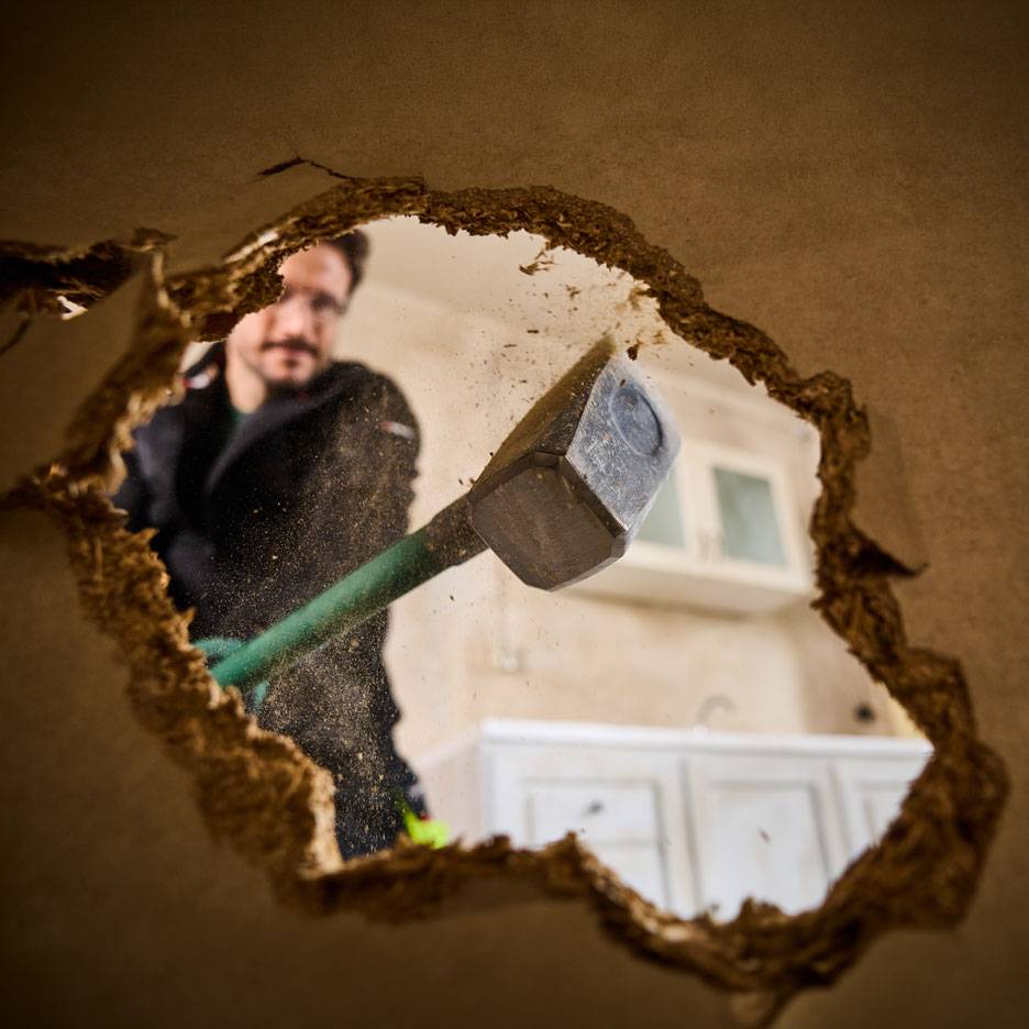 Muž s kladivom búra stenu, videný cez dieru, s kuchynskými skrinkami v pozadí.