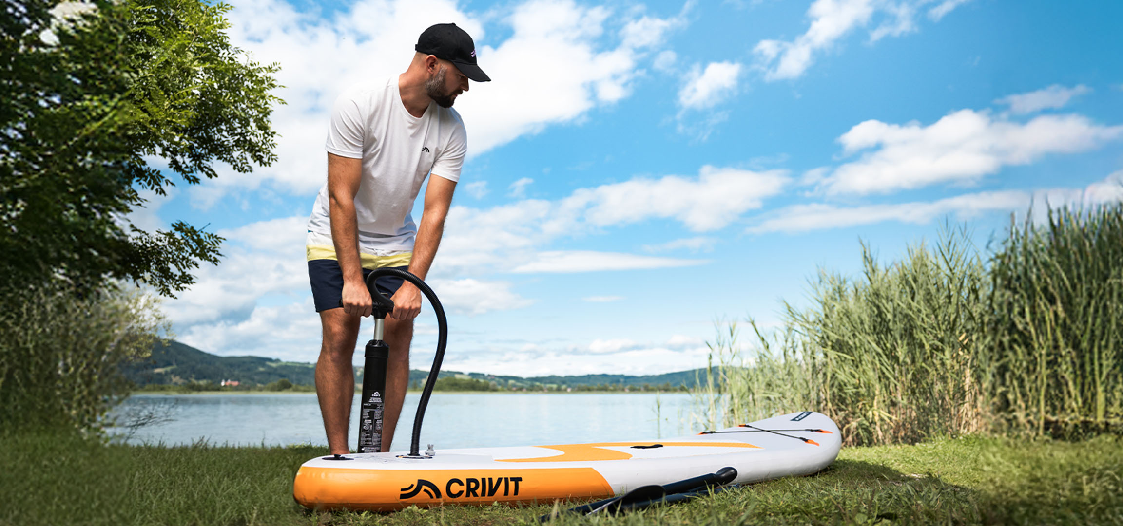 Muž nafukuje paddleboard CRIVIT pri jazere na zelenej tráve.