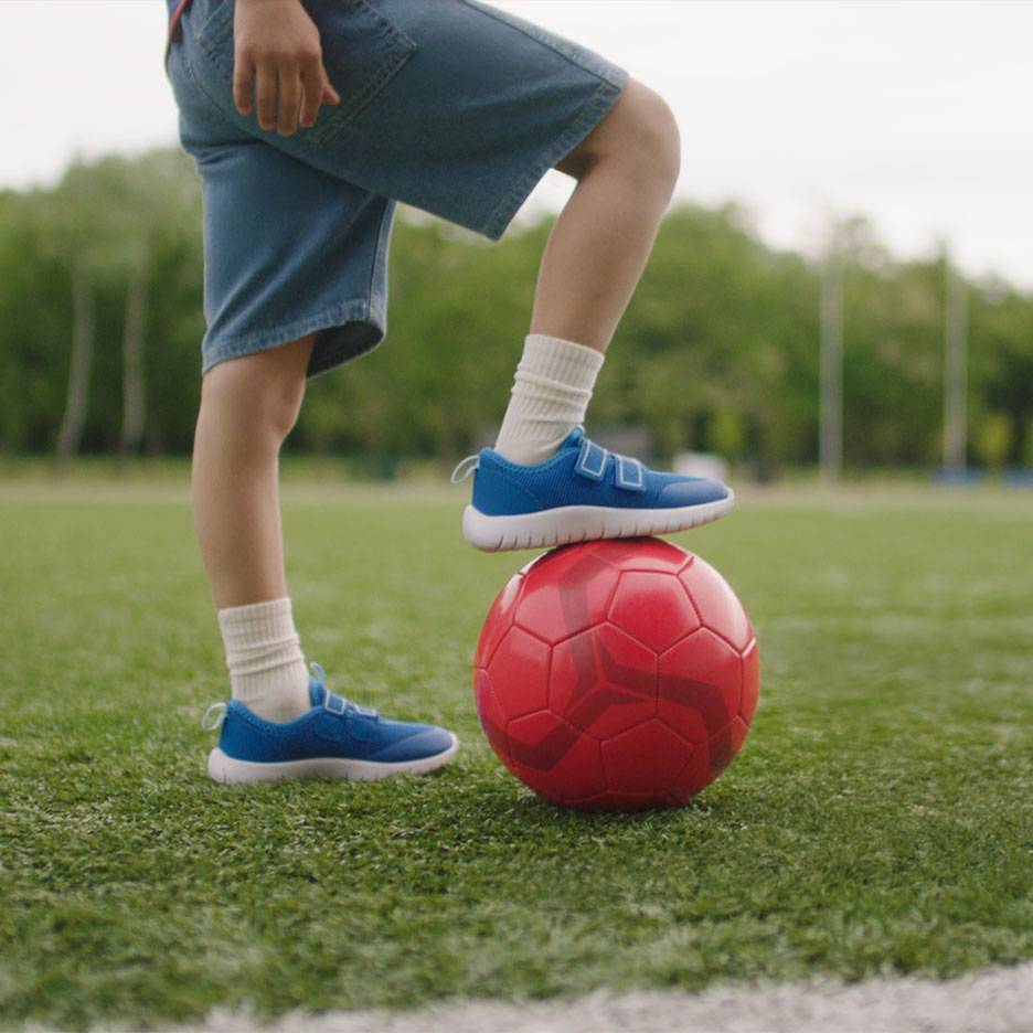 Dieťa v šortkách a modrých teniskách, s jednou nohou na červenej futbalovej lopte.