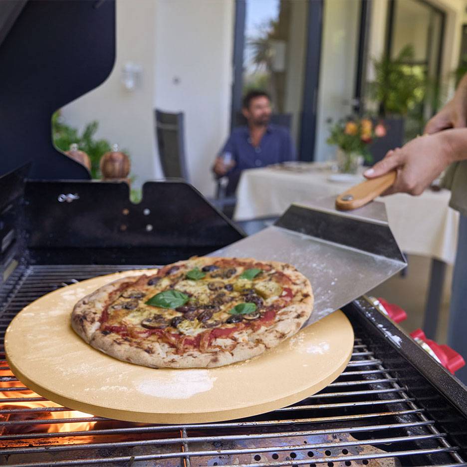 Pizza na pizza kameni na grile, s lopatkou na pizzu a mužom v pozadí.