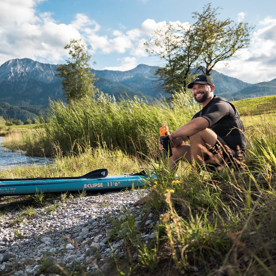 Usmievajúci sa muž v šiltovke a záchrannej veste, sediaci pri modrom paddleboarde pri rieke.