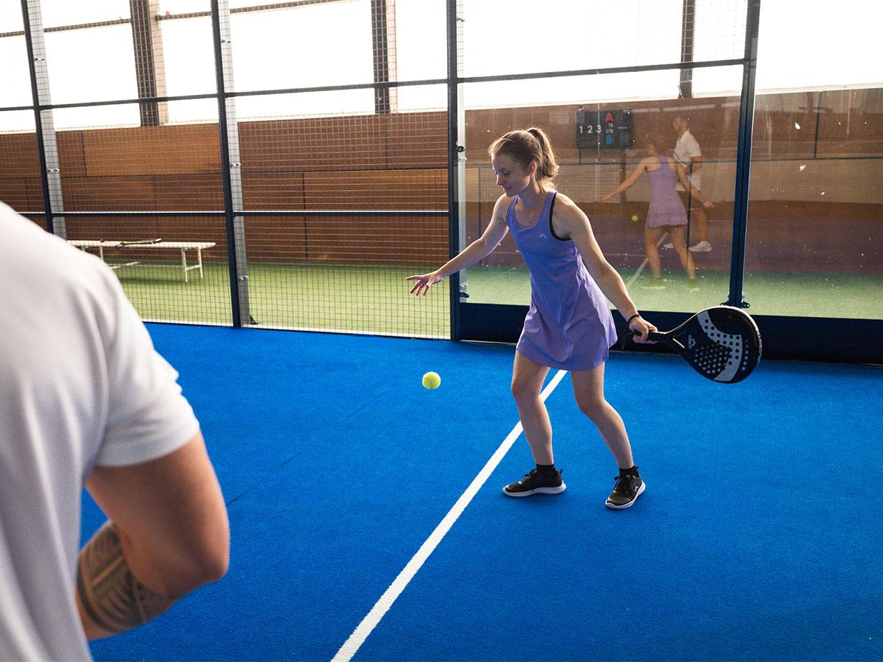 Žena vo fialových športových šatách hrá padel s raketou a loptičkou.