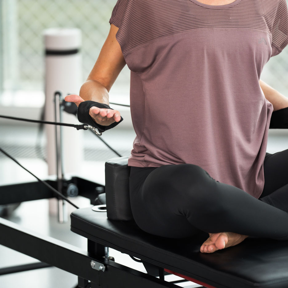 Žena cvičiaca Pilates na reformer stroji, v čiernych legínach a fialovom tope.
