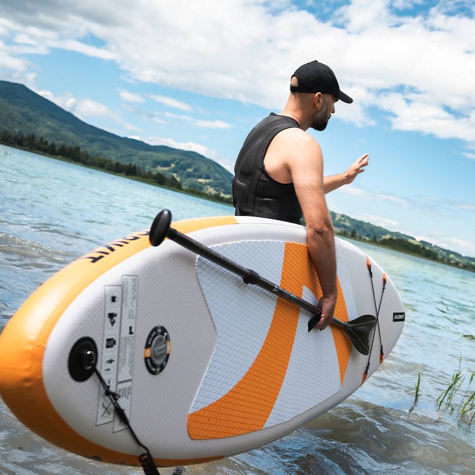 Muž v záchrannej veste a čiapke drží nafukovací paddleboard v jazere.