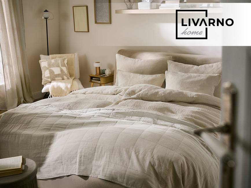 Spálňa s béžovou posteľnou bielizňou, vankúšmi a prešívanou prikrývkou, s nočným stolíkom a lampou.