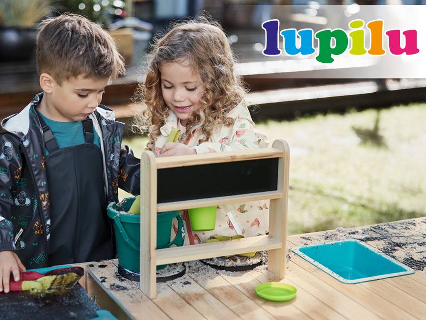 Dve deti sa hrajú s drevenou tabuľou a vedrom na pieskovisku.