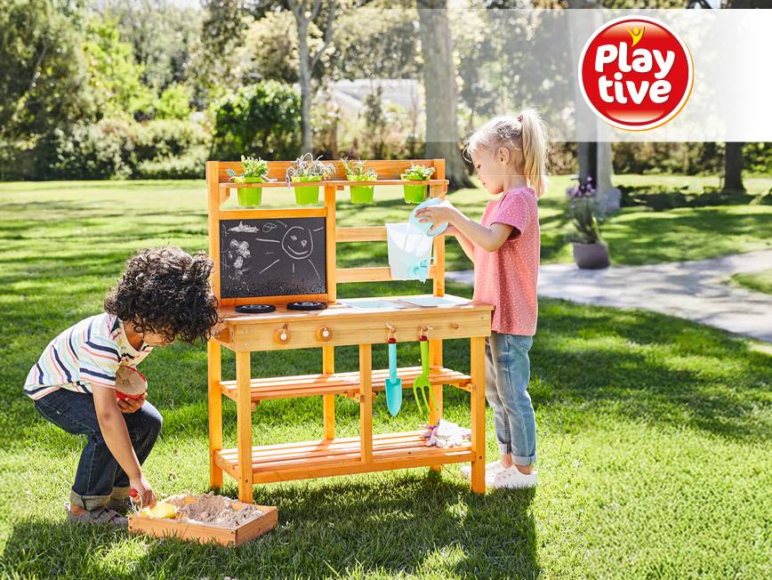 Dve deti sa hrajú s drevenou vonkajšou kuchynkou a pieskoviskom na zelenej tráve.