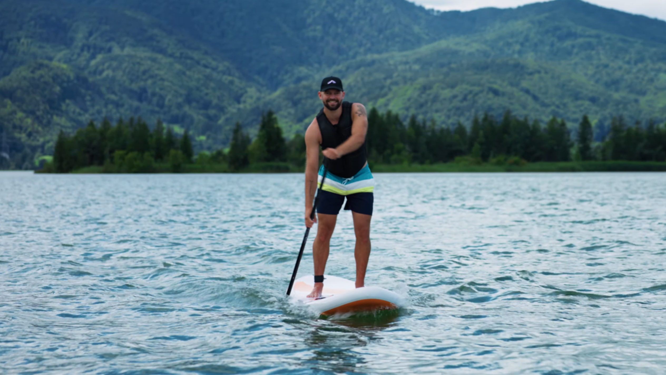 Muž na paddleboarde na jazere s horami v pozadí.